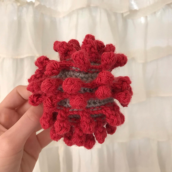 A photograph of a hand holding up the crocheted virus, showing us the back side of the project, red spikes.